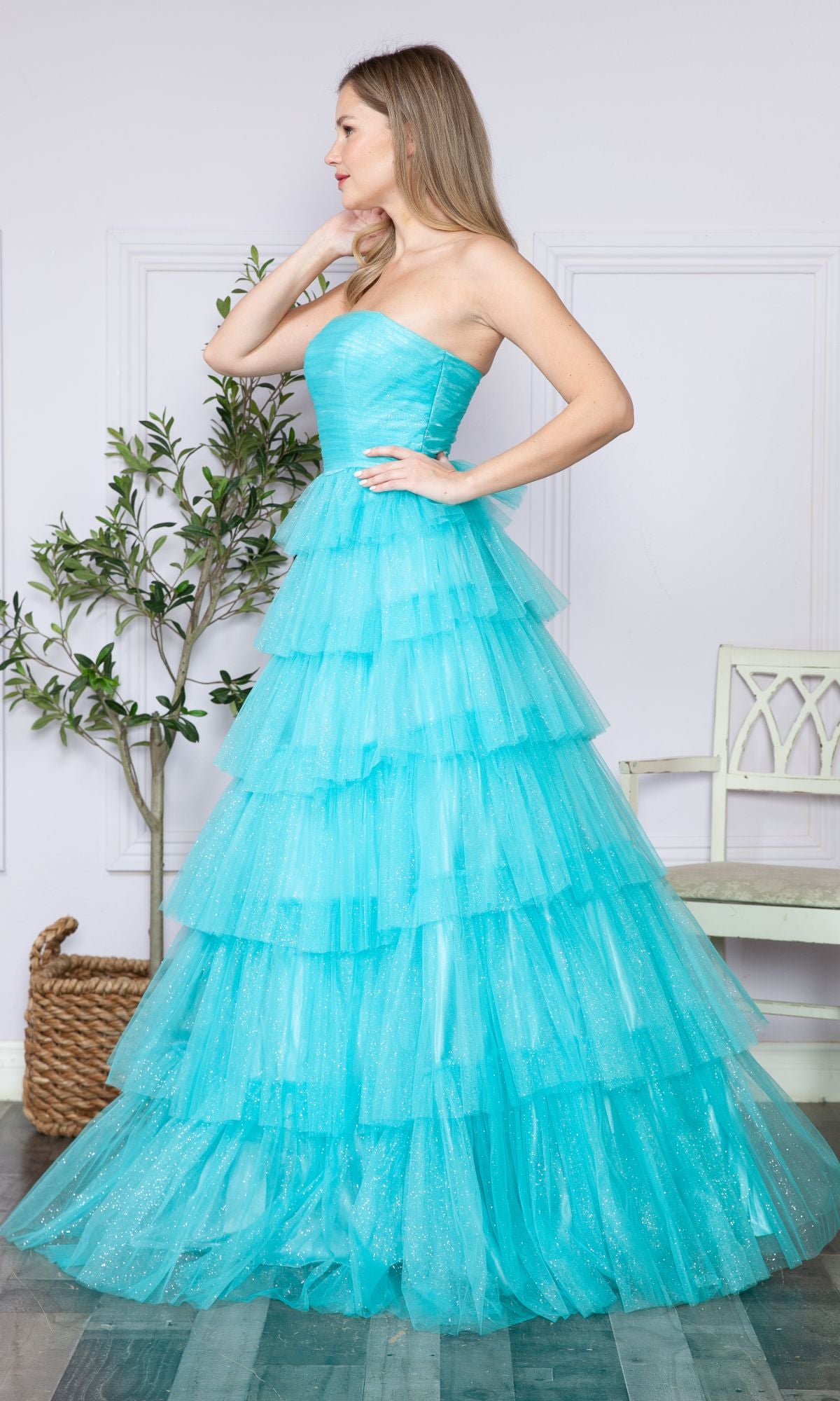 A woman stands indoors near a small tree and white chair, facing sideways, wearing the Strapless Long Tiered Glitter Prom Ball Gown 9386 in layered turquoise tulle.